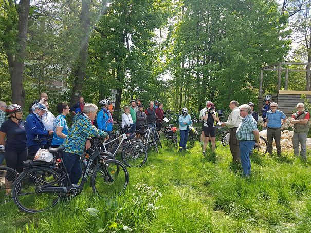 Viele Personen stehen mit oder auf Fahrrädern auf einer grünen Wiese. Im Hintergrund sind ein Holzgerüst und hohe Bäume zu sehen