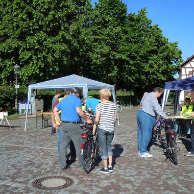 Zwei Gruppen stehen jeweils um ein Fahrrad und reden miteinander. Im Hintergrund stehen zwei Pavillons der Lokalen Agenda 21 Allensbach vor grünen Bäumen und einem Fachwerkhaus.