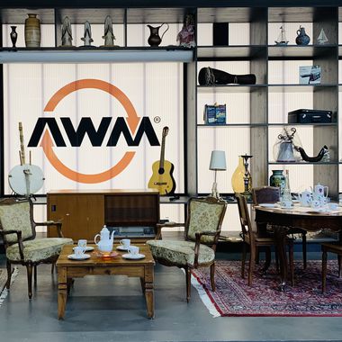 Vor einer beleuchteten Wand mit dem Logo "AWM" der Abfallwirtschaftsbetriebe München stehen Kaffeetische mit Stühlen in klassischem Stil mit gedeckten Teeservice. Im Regal stehen verschiedene Gegenstände wie Vasen, eine Gitarre oder Lampen.