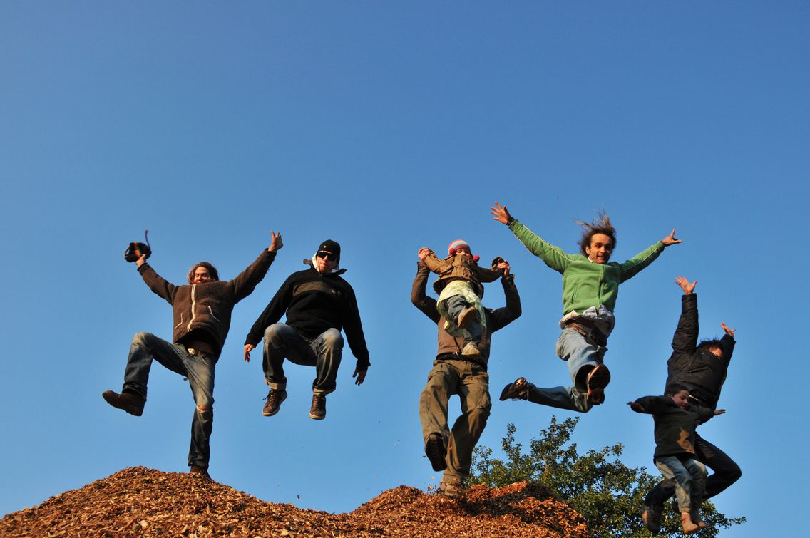 Sieben Mitglieder von Wirundjetzt e.V. springen gemeinsam für das Foto von einem Haufen Holzschnitzel herunter.