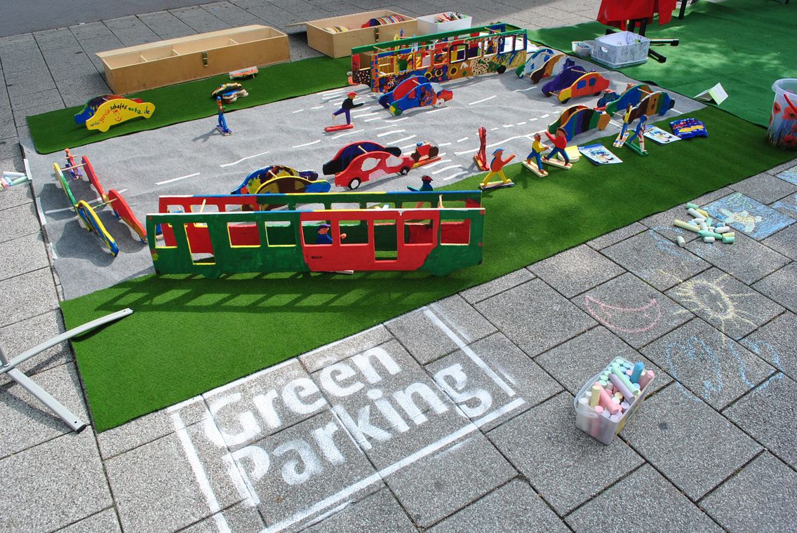 Eine Spielmatte mit Straßen und Grünstreifen auf der Verkehr simuliert werden kann mit bunt bemalten Autos, Bussen und Fußgängern aus Holz. NEben einem Eimer Kreide steht der Slogan "Green Parking Day".